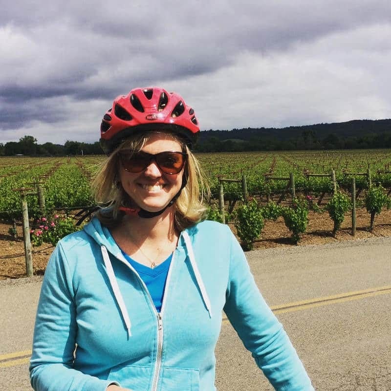 A woman wearing a helmet is standing in front of a vineyard.