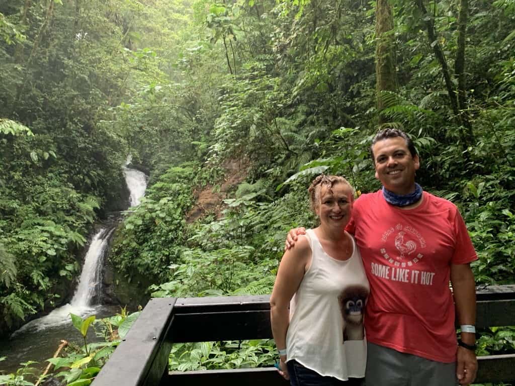 A man and woman standing in front of a waterfall in the jungle.